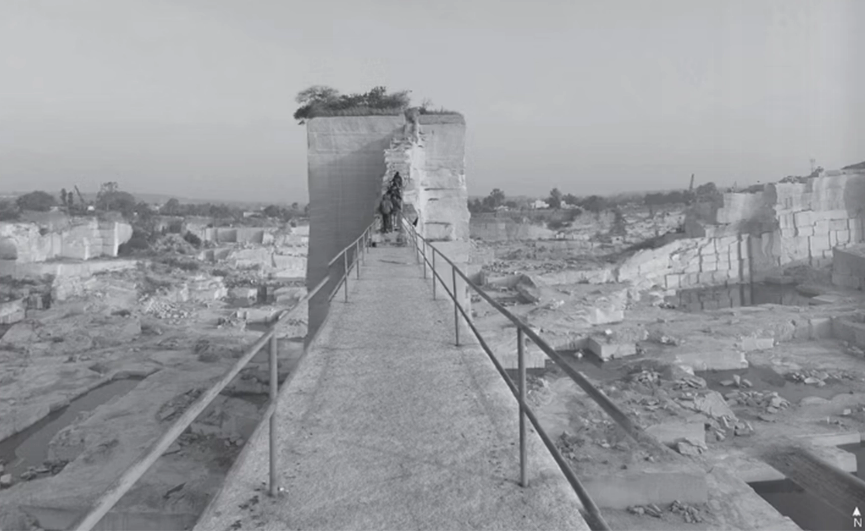 Existing quarry walkway approaching a solitary cut-stone tower across the excavated landscape.